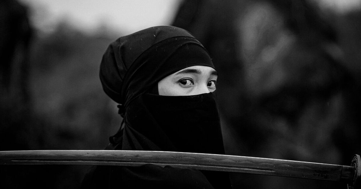 Black and white portrait of a woman in a hijab with a sword, gazing back.