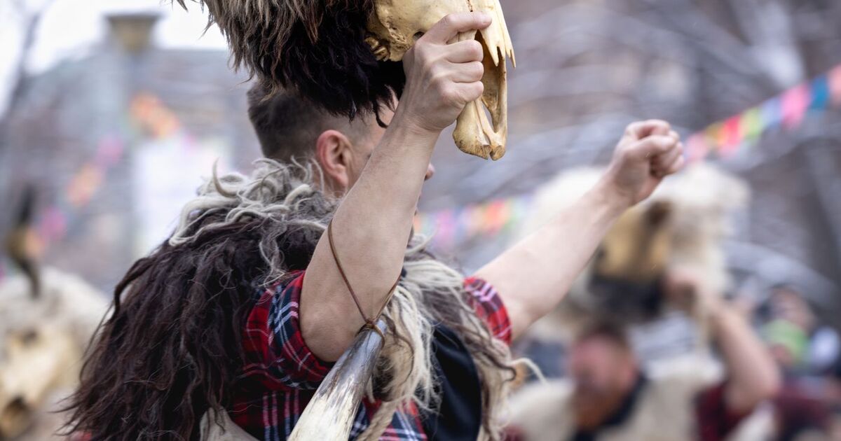 Man in furs and plaid shirt holding animal skull at an outdoor festival, embodying cultural traditions.