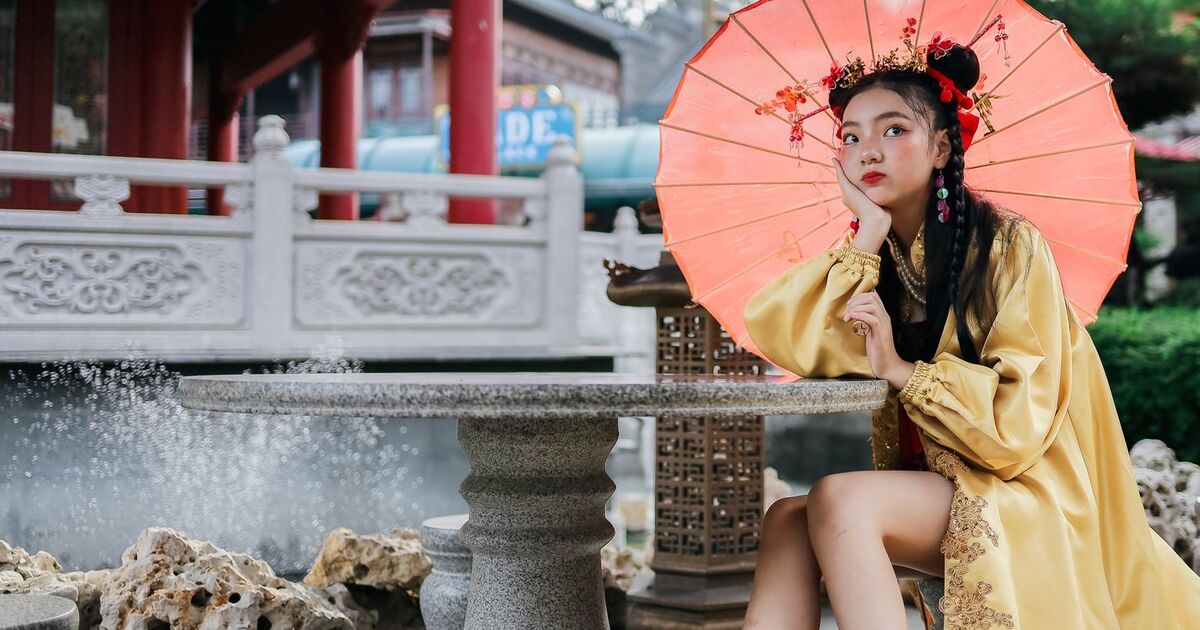 Woman wearing traditional Chinese attire sitting outdoors with a vibrant parasol.