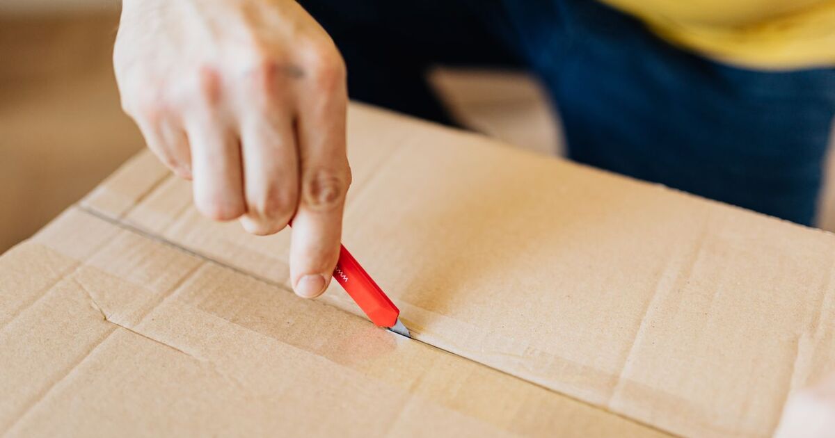 Crop anonymous male using utility knife to unseal packed cardboard box after receiving delivery during relocation