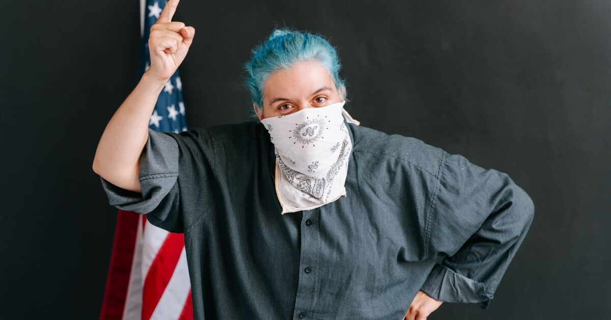 Person with blue hair and mask raises hand in protest beside American flag. Expressive activism.